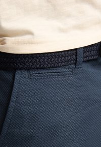 Camisa beige metida dentro de pantalones azules texturizados con un cinturón tejido en color azul marino oscuro pasado por las trabillas.