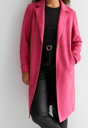 Femme portant un long manteau rose vif par-dessus un haut noir et un pantalon noir ceinturé, debout avec une main dans la poche de son manteau.