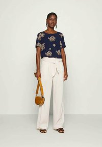 Mulher a usar uma blusa curta de manga com estampa floral em azul marinho, calças brancas de perna larga de cintura alta com cinto, sandálias castanhas e a segurar uma bolsa beje.