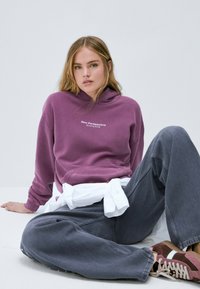 Un sweatshirt violet avec un logo blanc indiquant "New Perspective Browina 2006", associé à un pantalon gris ample et des baskets marron.