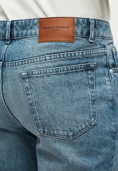 Πλάτη ελαφρώς γαλάζιων τζιν από τζίν με εμφανές καφέ δερμάτινο ετικέτα που γράφει "Officine Générale Paris 6" πάνω από την πίσω τσέπη.