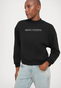 Čierna mikina vyrobená z bavlny, s rebrovanými manžetami a výstrihom, s logom "Armani Exchange" v bielej farbe pretekajúcom cez prednú časť.
