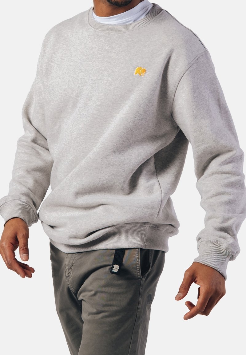 Homme portant un sweatshirt gris clair avec un petit logo d’éléphant jaune sur la poitrine et un pantalon gris, debout devant un fond uni.