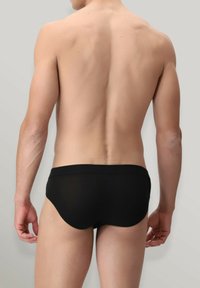 Slip da bagno neri con una texture liscia, caratterizzati da una vita bassa e una copertura minima, visti dal retro su uno sfondo neutro.