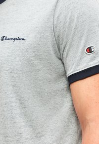 Γκρι μπλουζάκι Champion με σκούρο μπλε λεπτομέρεια στο μανίκι και κεντημένο λογότυπο Champion στο στήθος και το μανίκι, φορεμένο στο χέρι.