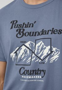Tekst na koszulce "Przekraczając Granice" z grafiką gór pokazującą wysokie i niskie szczyty, podpisane "Country Pacemakers S.Oliver Elevated Casual."