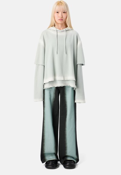 Jasnozielona, oversize bluza z kapturem o warstwowym wzorze, zestawiona z szerokimi spodniami w gradientzie czerni i turkusu. Gładka tekstura, minimalne detale.