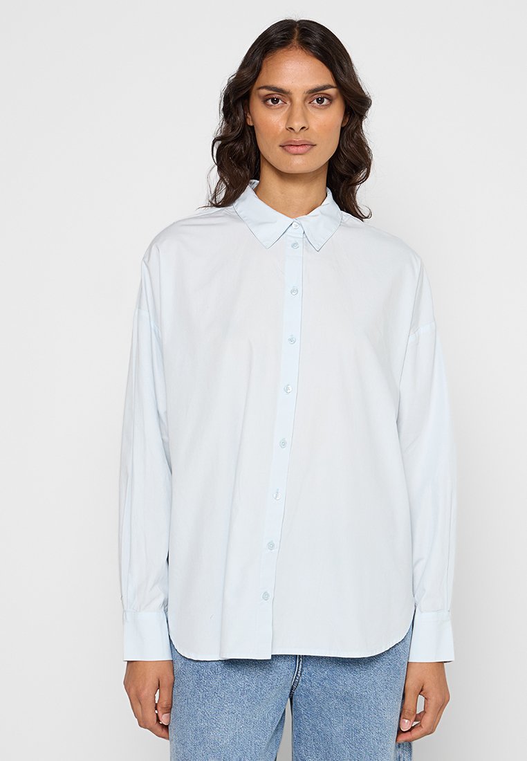 Donna con capelli scuri e mossi, che indossa una camicia button-up oversize di un blu chiaro e jeans di denim blu, in piedi contro uno sfondo neutro.