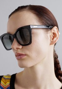 Czarne okulary przeciwsłoneczne z dużymi, kwadratowymi oprawkami i ciemnymi soczewkami; teksturowane detale na oprawce, z delikatnymi srebrnymi akcentami.