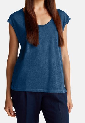 Femme aux longs cheveux bruns portant un haut bleu à manches courtes et un pantalon bleu foncé, sur un fond uni.