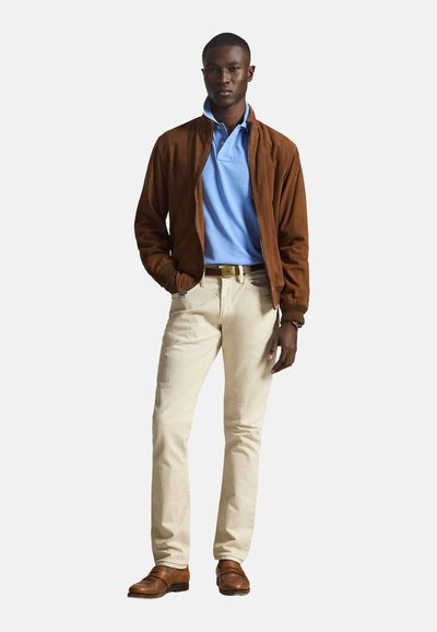 Homme portant une veste en daim marron, un polo bleu clair, un pantalon crème, une ceinture marron et des mocassins marron, debout avec une main dans la poche.