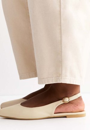 Füße in beigen Slingback-Flats mit goldenen Schnallen, kombiniert mit cremefarbenen, weit geschnittenen Hosen.