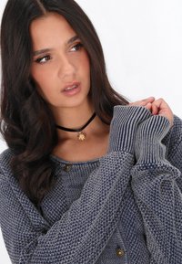 Cardigan grigio lavorato a maglia con polsini a coste, dotato di scollo rotondo e dettagli con bottoni dorati. Indossato con un choker nero e motivi testurizzati.
