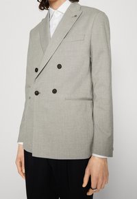 Šedý dvouřadý blazer z texturované tkaniny, se tmavými knoflíky, klopovými límečky a slim fit střihem, doplněný bílou košilí pod ním.