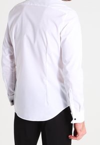 Mężczyzna ubrany w białą long-sleeve koszulę z czarnymi guzikami przy mankietach i czarne spodnie, pokazany od tyłu.