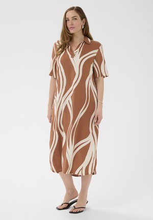 Kobieta ubrana w midi sukienkę w abstrakcyjny wzór w odcieniach brązu i kremu, z krótkimi rękawami, stojąca boso na białym tle.