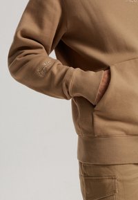 Persona con sudadera y pantalones beige, con una mano en el bolsillo de la sudadera, mostrando un texto bordado en la manga.