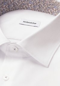 Witte shirt met een gestructureerde afwerking, voorzien van een gegarneerde kraag in lichtcrème, met een blauw en roze bloemenpatroon. Inclusief een enkele knoopsluiting.