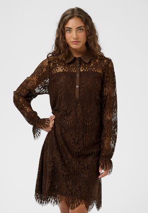 Femme aux cheveux bouclés portant une robe en dentelle marron foncé à manches longues avec un col et une patte boutonnée, debout avec une main sur la hanche.