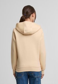 Sudadera beige con capucha ajustable, puños y dobladillo acanalados. Tela suave con una textura lisa. Combinada con jeans azules.