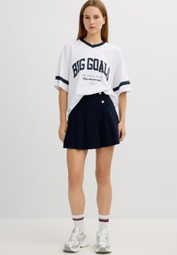 Camiseta blanca de gran tamaño con rayas azules y texto, combinada con una falda plisada azul marino con un detalle de botón, llevada con zapatillas blancas.