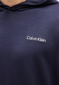 Μπλε σκούφο με το λευκό λογότυπο Calvin Klein κεντημένο στην αριστερή πλευρά του στήθους, φορεμένος από ένα άτομο.
