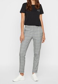 Kvinna iförd svart kortärmad skjorta med liten grafik och grå rutig slim-fit-byxa, kombinerat med vita sneakers, stående mot vit bakgrund.