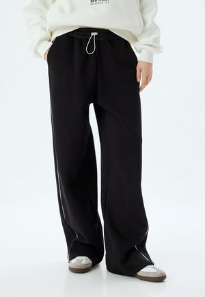 Pantalones de chándal negros con un diseño de pierna ancha, cinturilla elástica y cordón ajustable. Presentan un piping blanco en los laterales y una textura de tela suave.