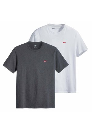 Mann mit lockigem Haar, der ein schlichtes weißes Kurzarm-T-Shirt und beige Hosen trägt, steht mit dem Rücken zu einem weißen Hintergrund.