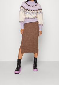 Kvinna i en lila mönstrad tröja, brun stickad midikjol med sidov slit och svarta ankelskor med lila platåsålar.