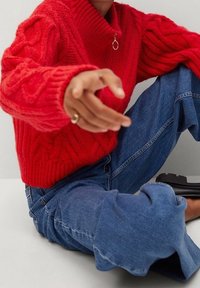 Pull en tricot rouge avec un col haut à côtes, featuring des motifs en torsades ; associé à un jean en denim bleu, affichant une coupe décontractée et des poignets effilochés.