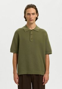 Jeune homme aux cheveux bruns mi-longs portant un polo en maille à manches courtes vert olive et un pantalon marron foncé, debout devant un fond blanc.