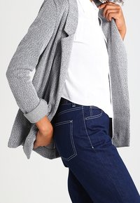 Šedý herringbone cardigan s volným střihem a ohrnutými rukávy, vrstvený přes bílou košili, spárovaný s tmavě modrými slim-fit džíny.