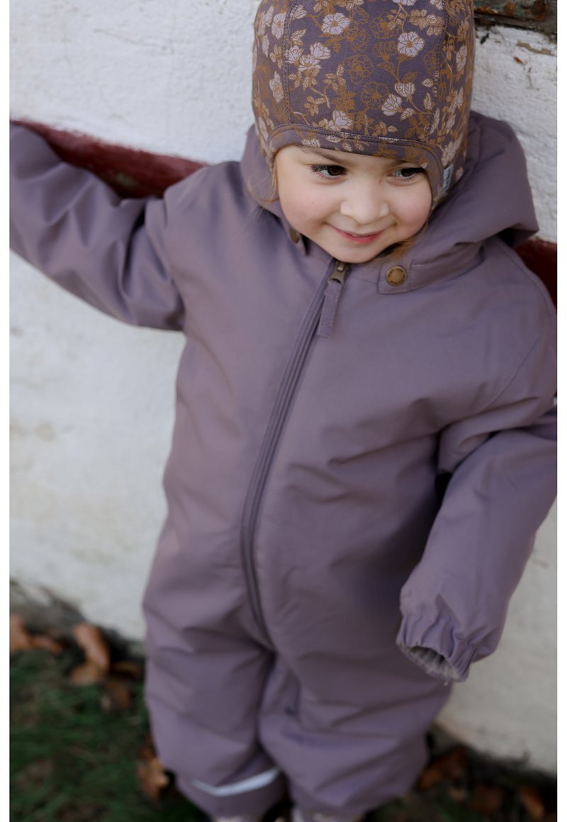 Fialová nepromokavá zimní kombinéza s kapucí, vybavená předním zipem, elastickými manžetami a hnědou čepicí s květinovým vzorem.