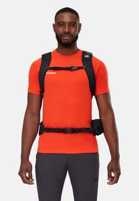 T-shirt de sport orange vif en tissu lisse, porté avec un sac à dos noir doté de bretelles réglables et de boucles pour un ajustement sécurisé.