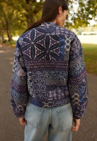 Chaqueta bomber acolchada en tonos de azul y púrpura, con varios patrones, mangas abullonadas y un dobladillo acanalado. Combinada con jeans de un azul claro.