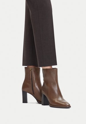 Braune Leder-Stiefeletten mit Blockabsätzen und seitlichen Reißverschlüssen, getragen zu dunklen, gekürzten Hosen auf hellem Hintergrund.