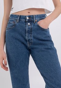 Denim bikses vidējā zila tonī, ar augstu jostu, divām priekšējām kabatām un aizdari ar divām pogām. Klasisks taisnās kājas dizains.
