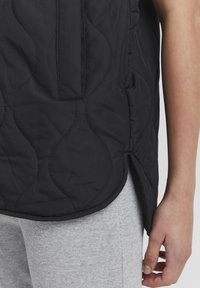 Schwarze gesteppte Weste mit geschwungenen Nähten und seitlichen Schlitzen, kombiniert mit grauen gerippten Leggings. Der Stoff wirkt glatt und leicht.