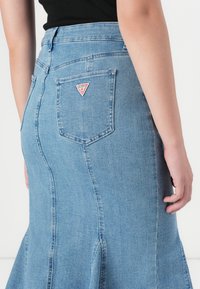 Een persoon in een zwart topje staat met de rug naar de camera en toont een blauwe denim rok met een zichtbaar merklogo op de zak.