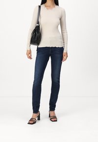 Top de manga larga beige de canalé con bordes de encaje en forma de ola, combinado con jeans slim de color azul oscuro y sandalias de tacón negro con tiras. Bolso negro.