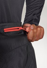 Pantalons de sport noirs en tissu texturé ; dotés d'une poche zippée orange à la taille et d'un gros plan d'une main tenant la tirette du zip.