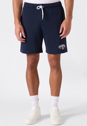 Männliche Beine, die marineblaue Shorts mit Kordelzug und Champion-Logo tragen, weiße Sportsocken und weiße Sneaker vor einfachem Hintergrund.