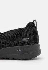 Sneaker en mesh noir avec un dessus texturé, une semelle en caoutchouc épaisse et un petit logo sur le côté. Onglet de traction doux à l'arrière pour un enfilage facile.