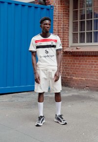 Maglia atletica bianca con strisce rosse e nere; logo "Snapdragon"; abbinata a pantaloni corti bianchi e scarpe da ginnastica nere con dettagli grigi.