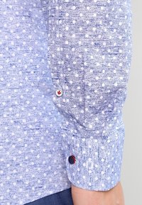 Light blue long-sleeve shirt with white polka dots, textured fabric, and button cuffs featuring a red and blue button detail.