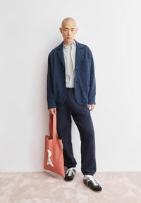 Marineblauwe katoenen blazer, licht geruit overhemd, marineblauwe broek en zwarte sneakers. Gecombineerd met een roze totebag met een wit grafisch ontwerp.