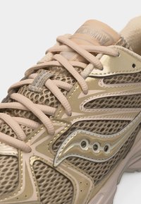 Μπεζ και χρυσό παπούτσι για τρέξιμο Saucony με επάνω μέρος από δίχτυ και μπεζ κορδόνια δεμένα πάνω από τη γλώσσα, με ορατό το λογότυπο της μάρκας.