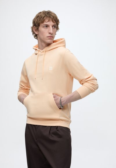 Jeune homme portant un sweat à capuche pêche clair et un pantalon foncé, les mains dans la poche avant, regardant sur le côté sur un fond uni clair.
