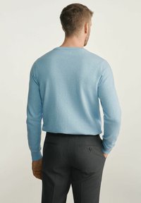 Mężczyzna odwrócony tyłem, ubrany w jasnoniebieski sweter i ciemnoszare dopasowane spodnie, z rękami w kieszeniach, na neutralnym tle.